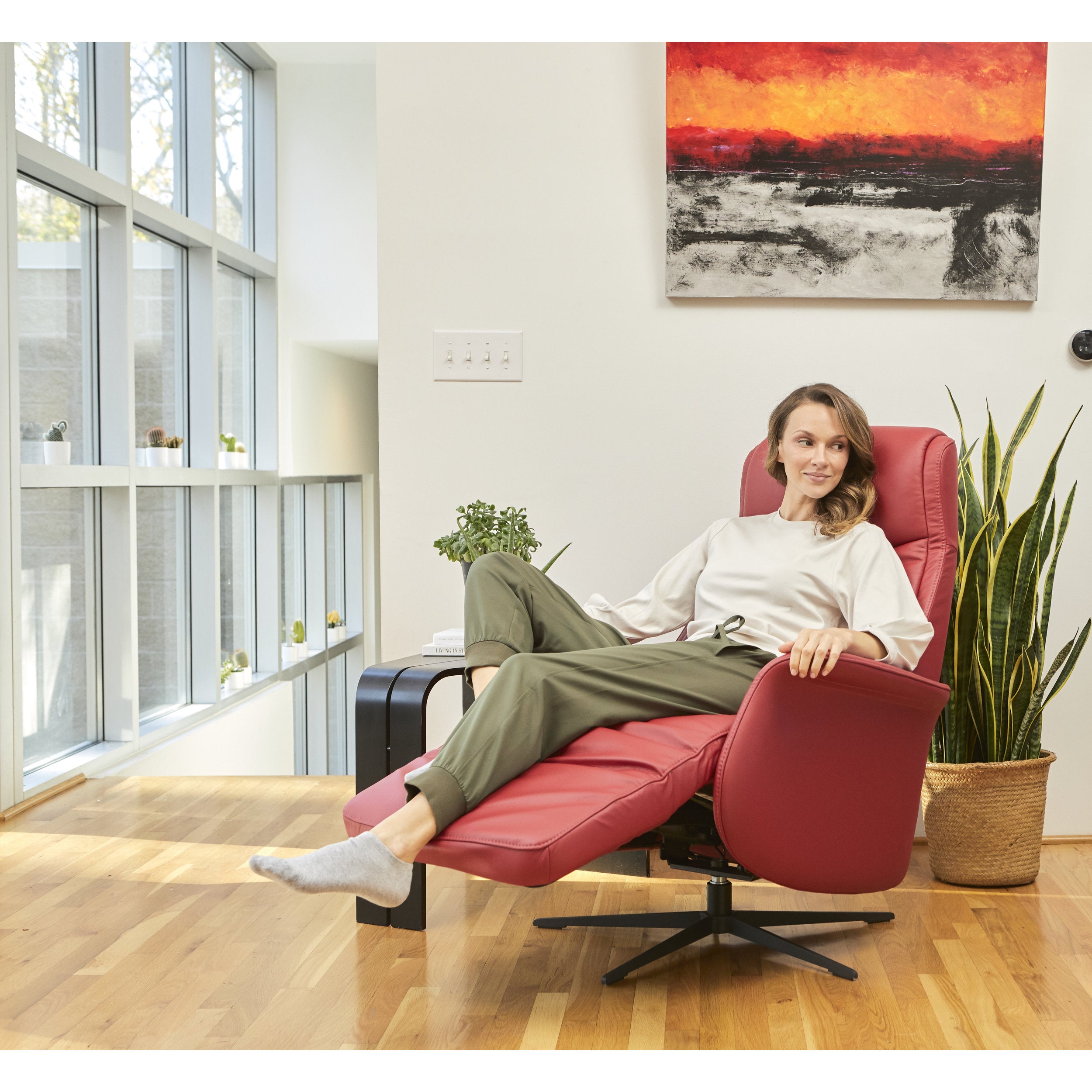 Woman relaxing in a red armchair in a modern living room with large windows and a painting on the wall.