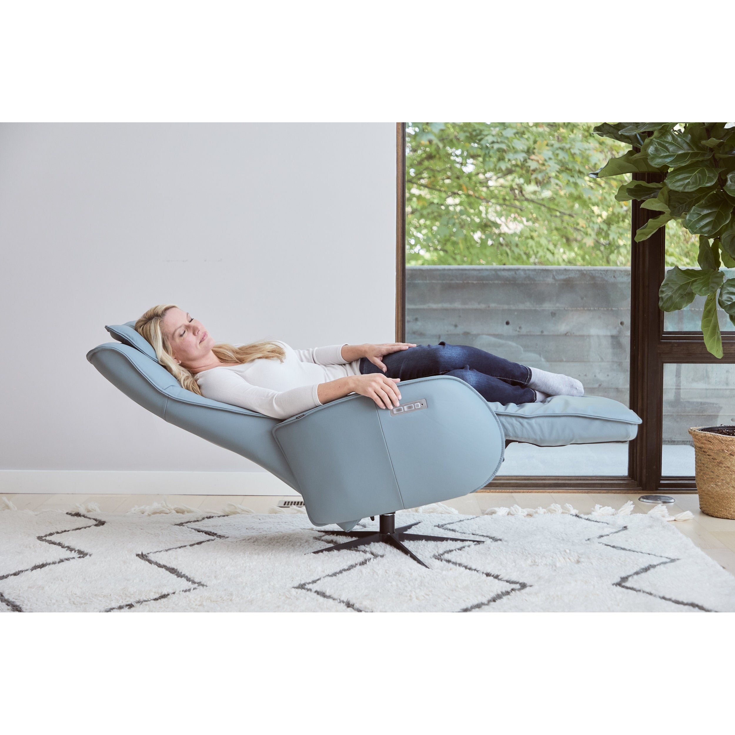 Woman relaxing in a modern blue chair in a room with a large window and plant.