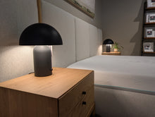 Load image into Gallery viewer, Modern bedroom with a black lamp on a wooden nightstand and a bed in the background.
