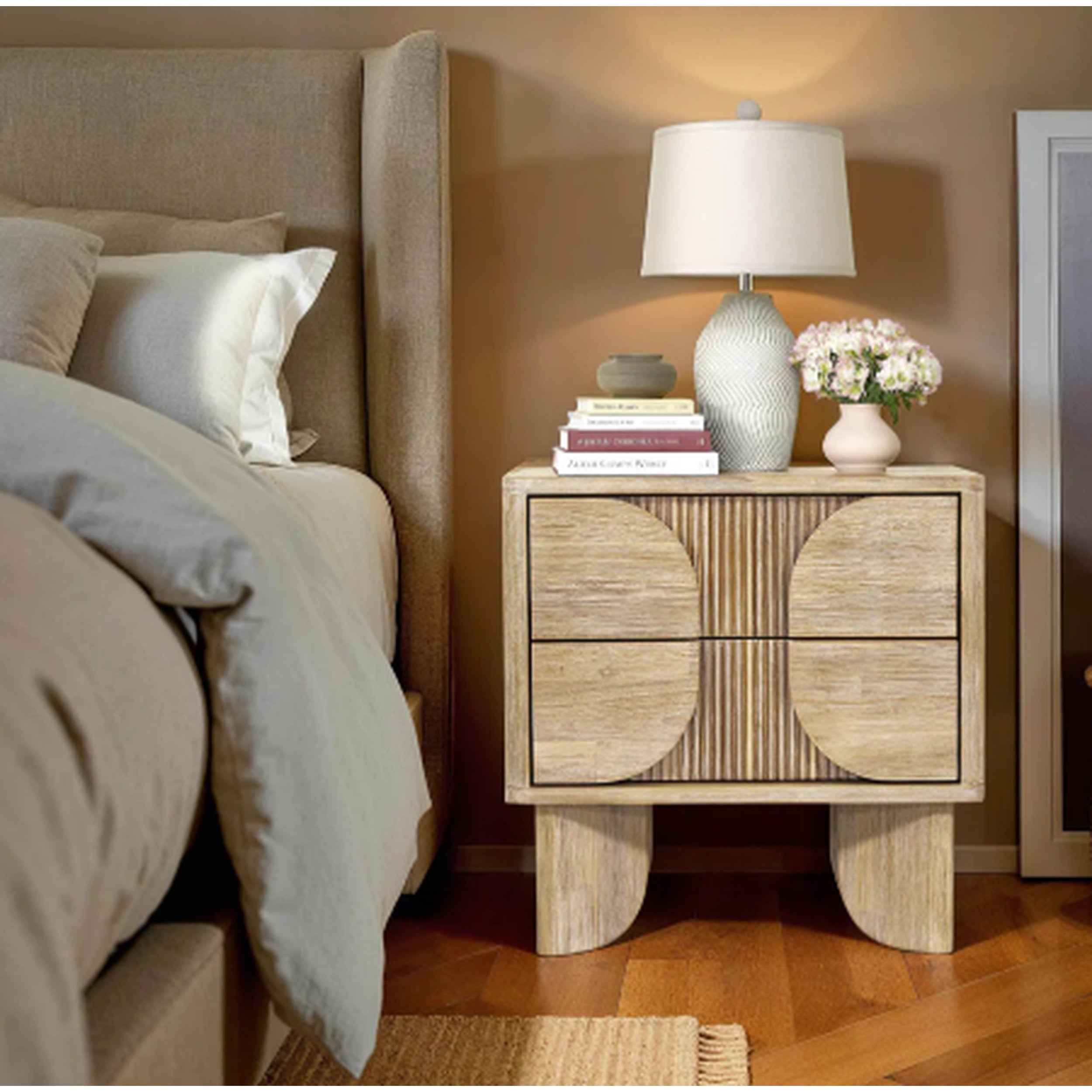 Wooden nightstand with lamp and books next to a bed in a bedroom setting